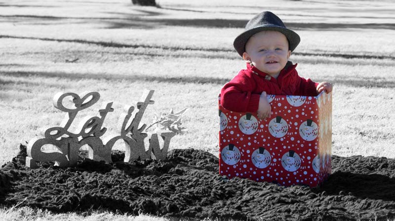 infant boy in christmas attire and fedora seated in box covered in wrapping paper outdoors on blanket next to let it snow sign for family diy holiday photos in mississippi by danielle jacqueline photography