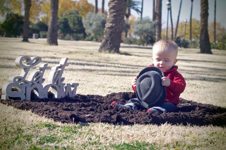 little boy sitting on brown blanket at park next to "let it snow" holiday decoration taking hat off cute photos by danielle jacqueline in olive branch ms