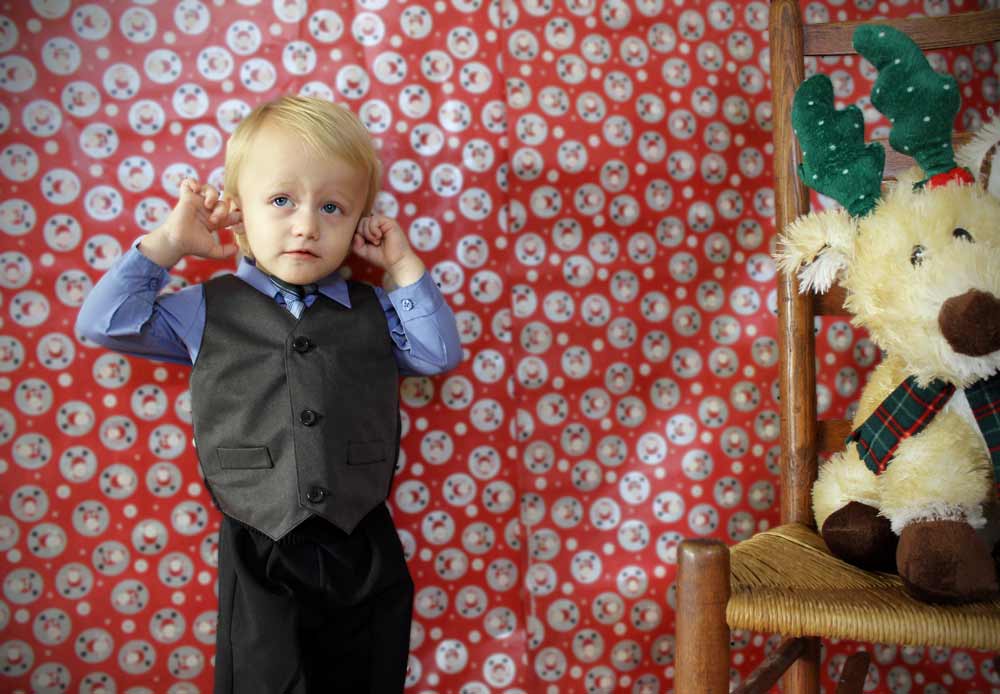 boy plugging his ears standing in homemade photography studio by danielle jacqueline south east of memphis tennessee with stuffed reindeer on wicker rocking chair and christmas wrapping paper background