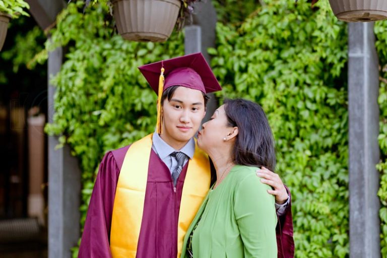 green sweater over dress mother leaning to kiss son on cheek as he looks at camera wearing cap and gown for college graduation by danielle jacqueline bright colored bushes in back