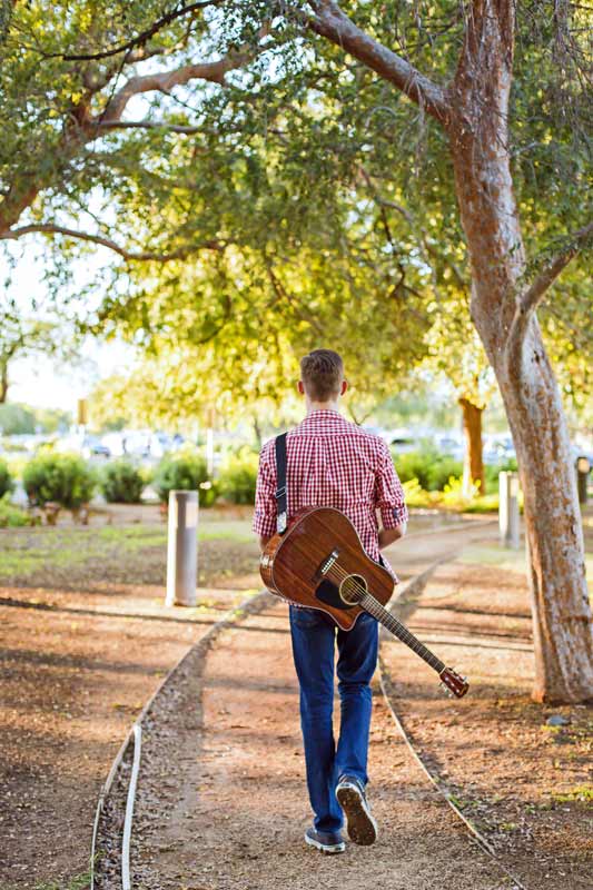 checkered white and red shirt with guitar slung over back of high school senior boy walking down path in autumn as leaves fall by mississippi photographer danielle jacqueline