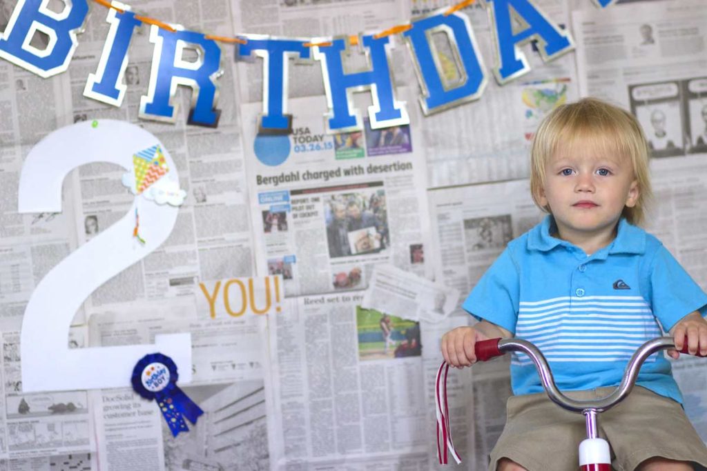 seated on tricycle uncomfortable face talking to photographer in olive branch ms who specializes in young kids at indoor studio blue theme with today's newspaper in background to signify date ideas