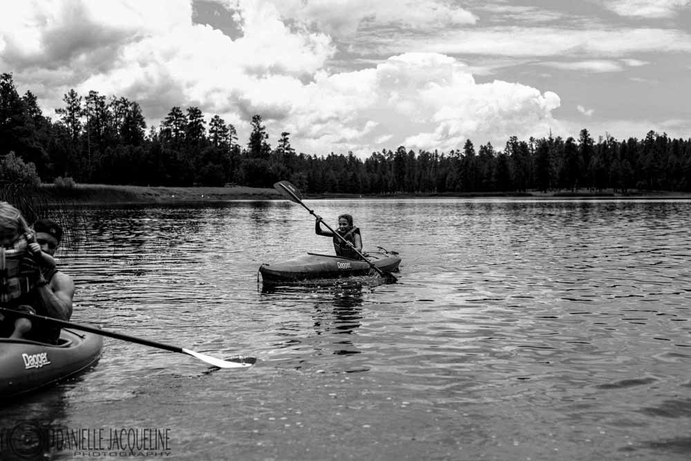 black and white family kayak trip on mississippi lake daughter behind in focus in water with tree line silhouette by danielle jacqueline photography in desoto county