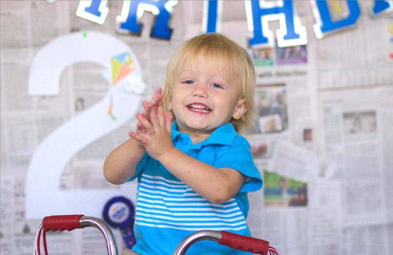 clapping little boy in baby blue long blonde hair smiling in front of two year old birthday celebration display behind him on bike by danielle jacqueline photography in olive branch ms