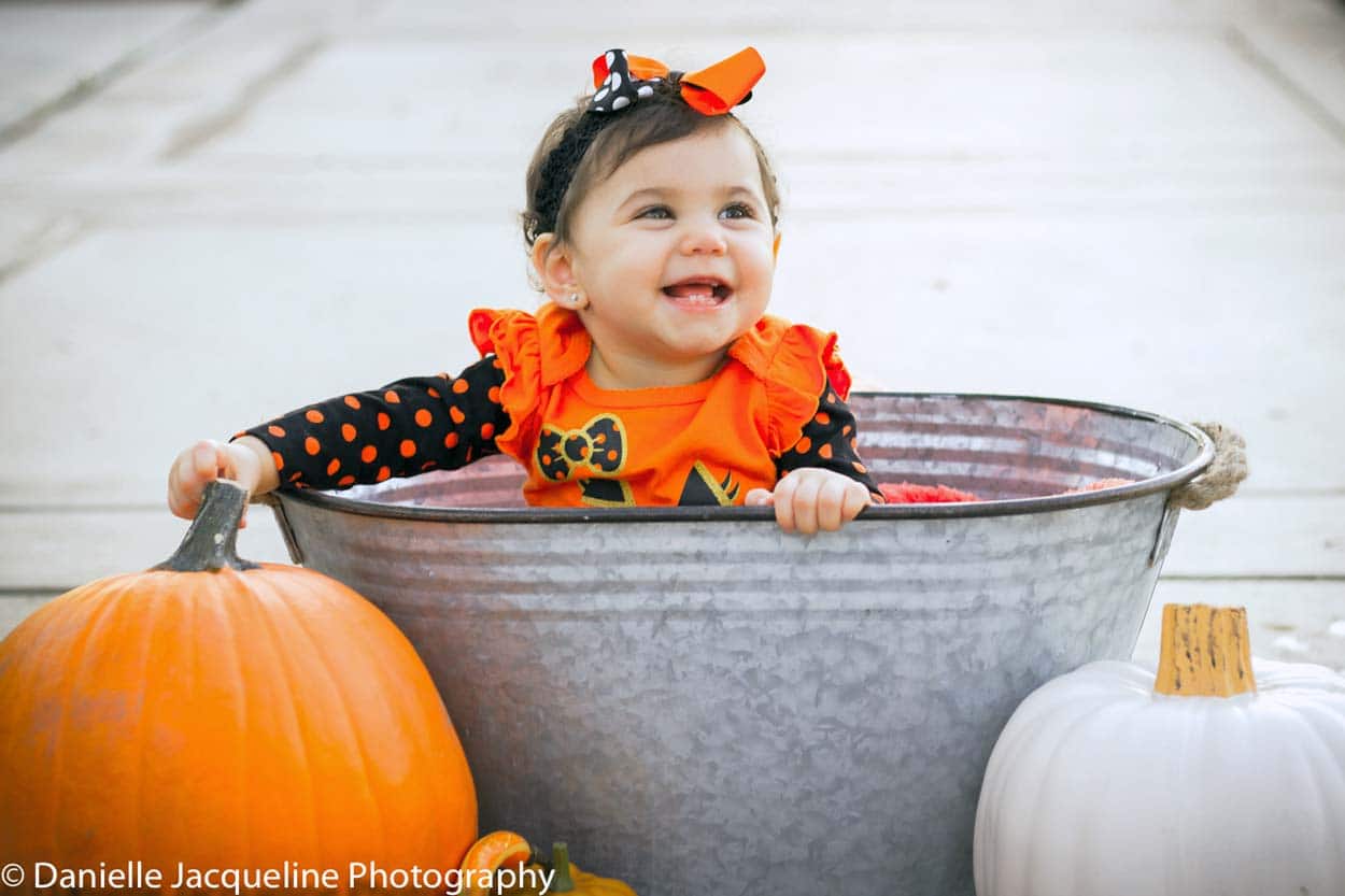 laughin baby girl looking to the right at mommy and daddy on sidewalk sitting in aluminum trough next to white and orange pumpkins enjoying the fall photography session with danielle jacqueline in desoto county ms