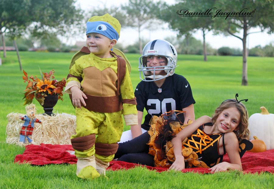 little boy in halloween costume walking away from sisters on blanket at park with pumpkins for fall photography ideas with kids natural lighting in olive branch ms with danielle jacqueline