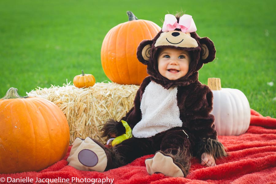 first halloween costume innocent little girl monkey suit onesie with hood surrounded by pumpkins on red blanket at the park in grass by danielle jacqueline photography in olive branch ms