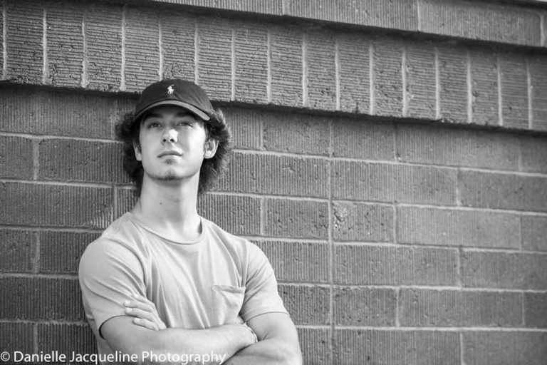 teen boy leaning back against brick wall posing arms crossed looking left ball cap black and white high school senior portrait by danielle jacqueline in olive branch ms