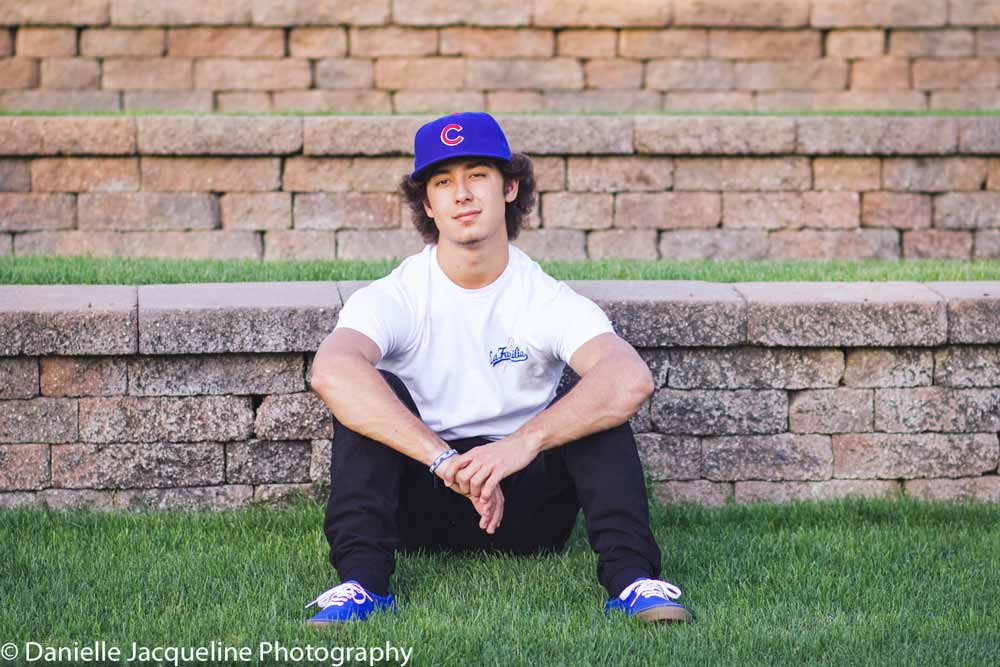 arms resting on knees sitting back against stone background with grass layers for senior portraits session with danielle jacqueline photography at dusk in olive branch ms