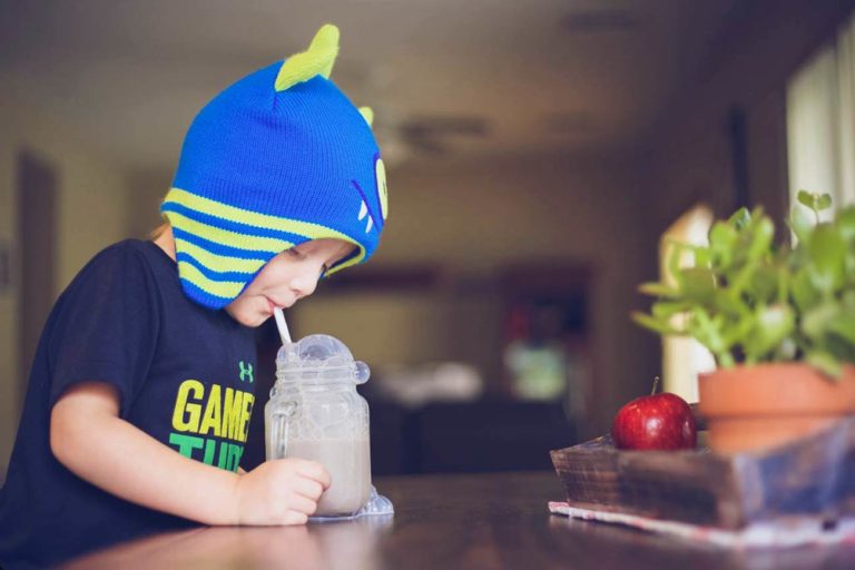 little boy monsters inc stocking cap blowing into straw glass jar chocolate milk bubbles overflowing half smirk at kitchen table with placement settings by olive branch photographer danielle jacqueline at home mississippi