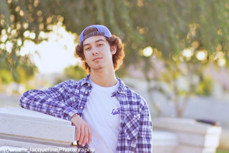 leaning against white picket fence line in front of fruit trees in blue flannel with backwards baseball hat arm up smirk at camera for senior pictures by danielle jacqueline in olive branch ms