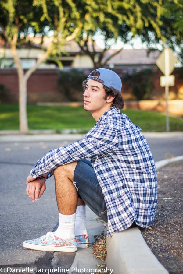 laid back senior graduate sitting on curb of neighborhood street looking off thinking backwards hat west coast style shoes and cut off jeans by danielle jacqueline photography in olive branch ms