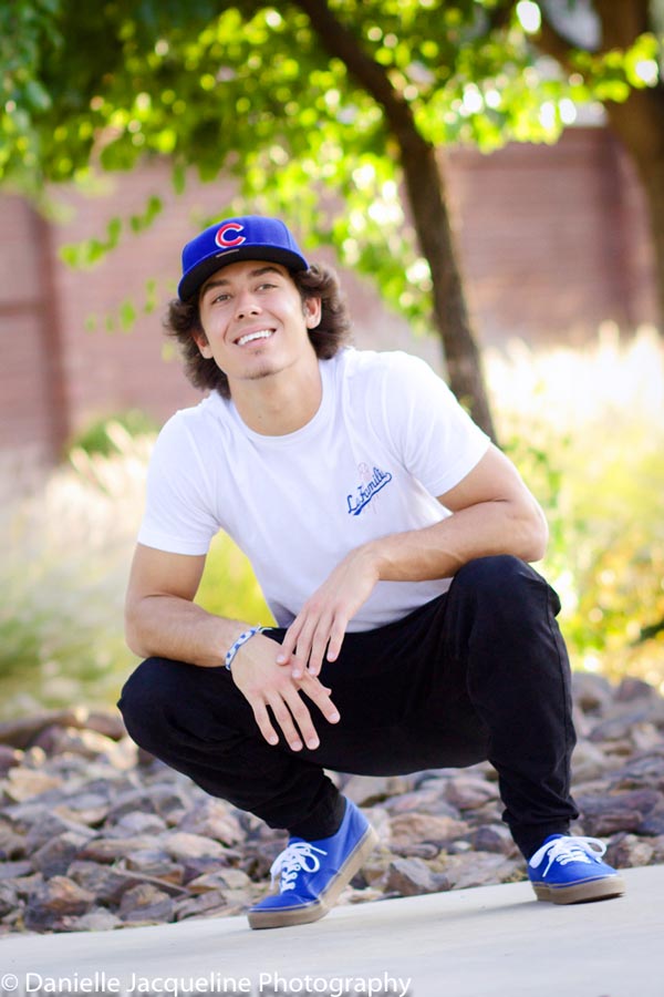 crouched down senior high school momma's boy white t shirt and jeans with blue cap and vans shoes on sidewalk spring day angled lens by danielle jacqueline photography in desoto county ms