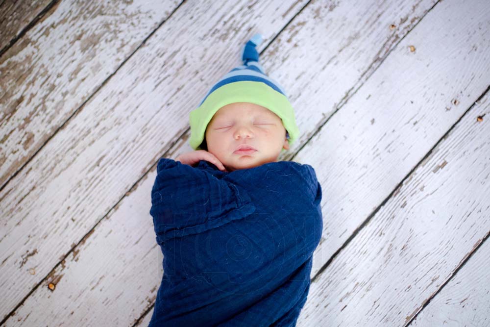 little boy born 10 days prior hand under chin wearing green and blue beanie with matching swaddle laying on back white wood flooring photography studio options for infants near memphis tennessee