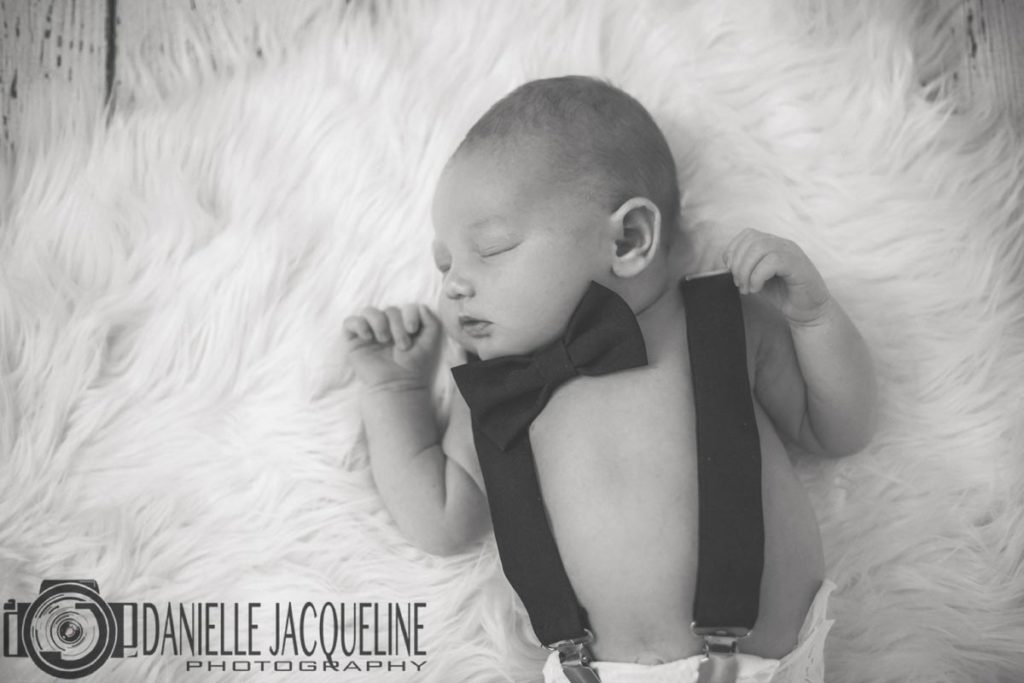 arms up baby boy newly born 10 days ago wearing suspenders attached to diaper black bow tie head to left sleeping on white fur rug at olive branch ms photography studio with danielle jacqueline