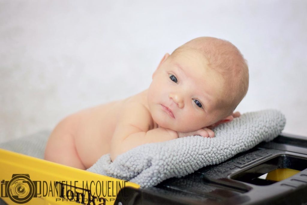 wide eyed fresh born baby portrait studio toy truck yellow in dump on blankie facing camera close with white decorated background by danielle jacqueline photography in desoto county ms