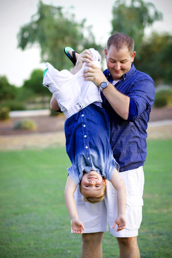 father in white shorts matching son holding little boy by legs with both hands at park with open green grass neutral background for summer family portraits new city midsouth mississippi photographer danielle jacqueline