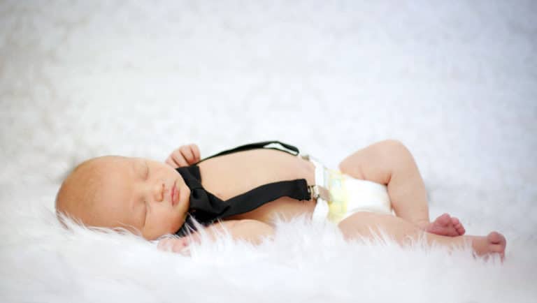 baby boy laying on back asleep during new arrival photo session with danielle jacqueline portrait studio in olive branch mississippi wearing diaper suspenders formal look on soft blanket white setting