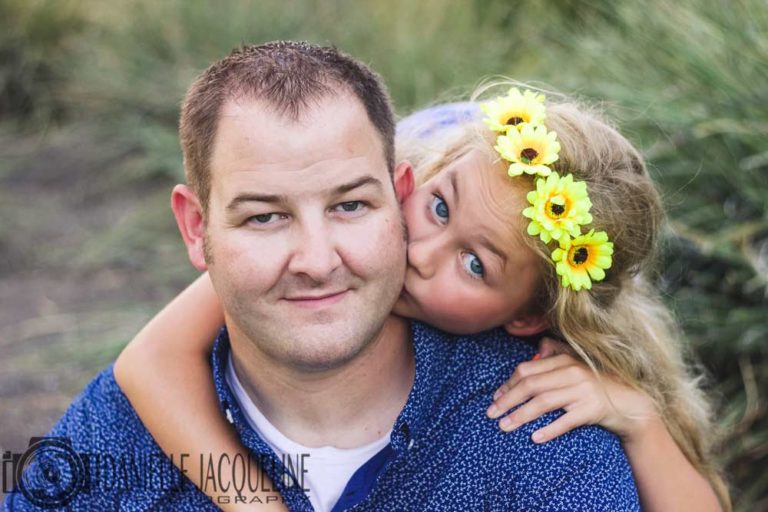 happy father looking up directly at camera lens with daughter flowers in hair giving him kiss on cheek hand on shoulder for family pictures during midsouth summer by danielle jacqueline photography