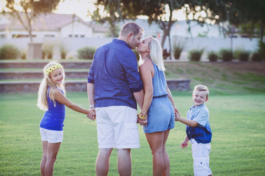 mom and dad kissing in the center at park green grass sunset rays with daughter on left smiling and son on right holding hands cheesing for summertime portraits for families with danielle jacqueline photography in olive branch ms