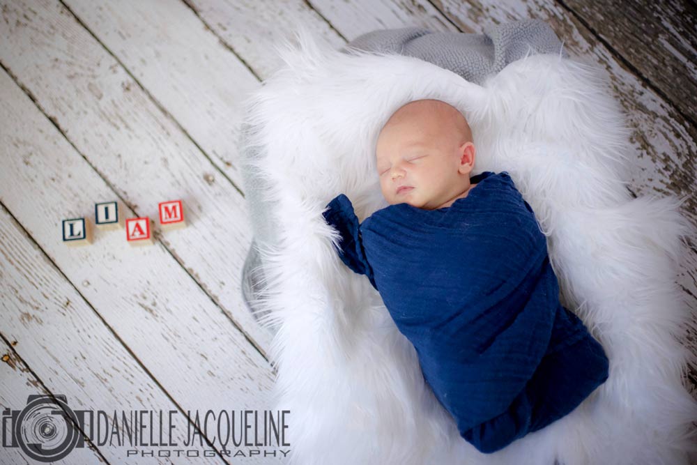 new arrival photographer for boys with son of happy parents laying on soft white blanket and wrapped in blue with name of child in blocks to the left on floor at danielle jacqueline portrait studio near memphis tn