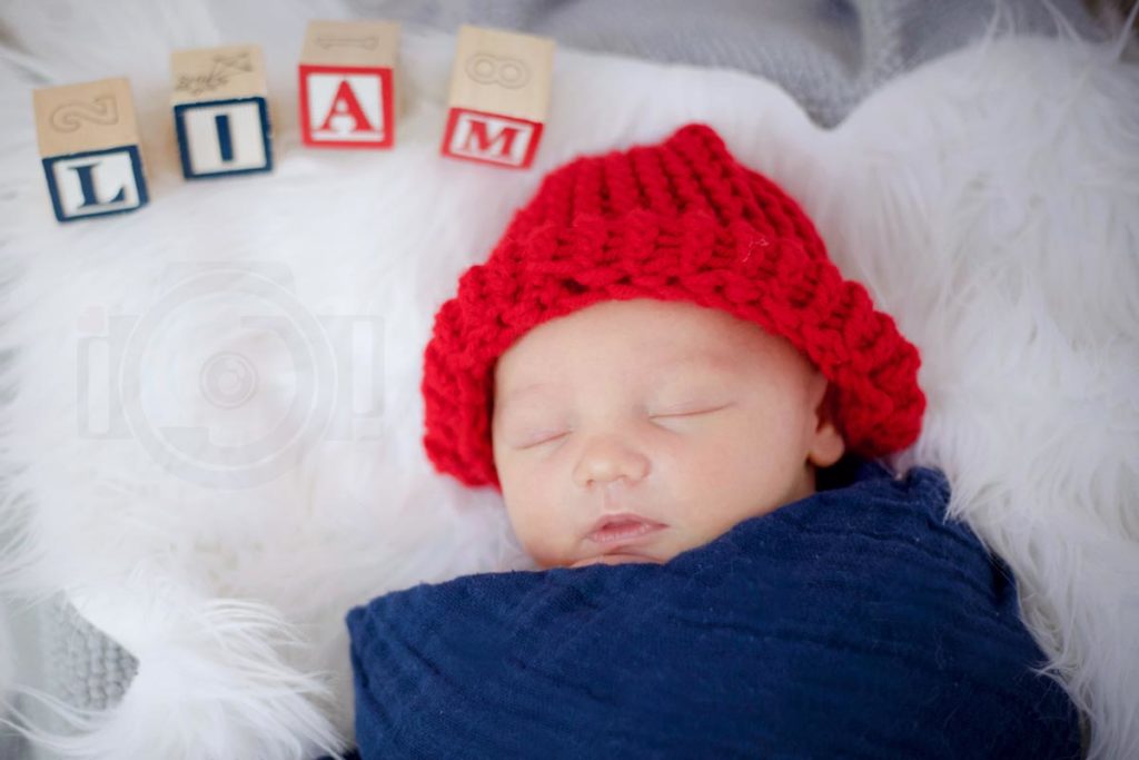 overhead close up shot of 10 day old baby boy wearing bright red beanie and dark blue swaddling with name in building blocks spelled out above head at danielle jacqueline portrait studio in olive branch mississippi