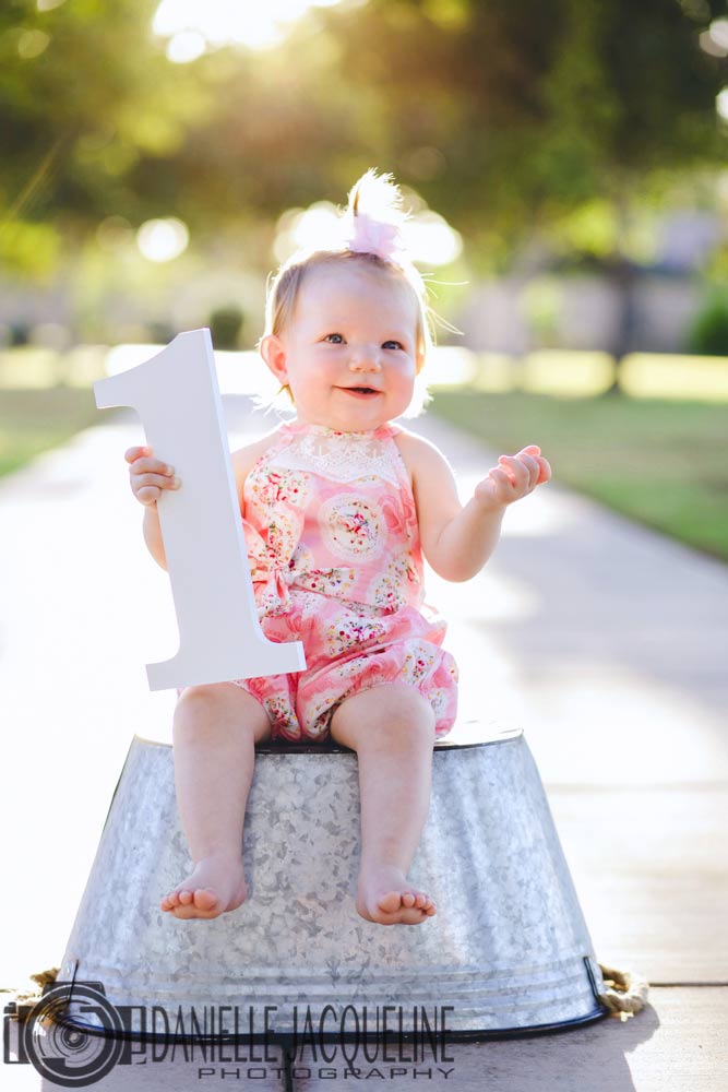 infant daughter pictures sitting on metal bin on sidewalk at park holding big number one cut out of wood for first birthday with danielle jacqueline in memphis tn