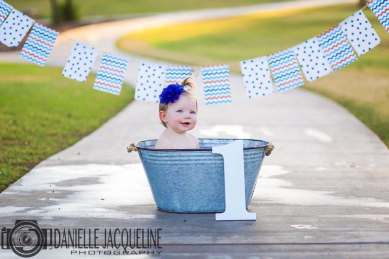 adorable one year old baby girl sitting inside metal tub with big number 1 and banner above for birthday pictures in olive branch by danielle jacqueline photography smiling