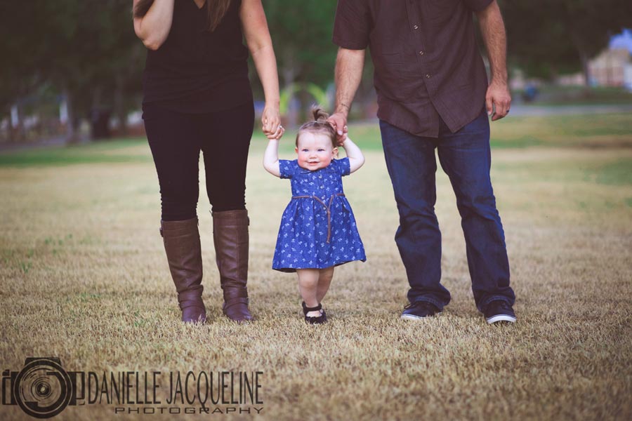 infant girl in blue polkadot dress walking towards camera in grass hand in hand with mommy and daddy for 1 year old birthday pictures in south memphis with danielle jacqueline photography