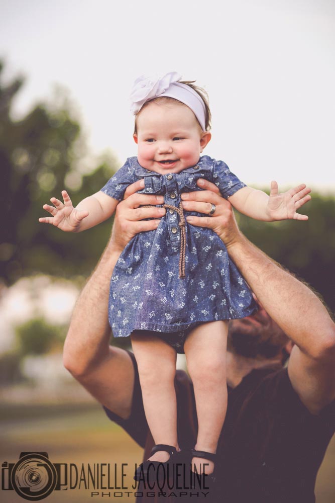 cheerful baby girl in blue dotted dress with boy arms up as daddy lifts her into the air during session with danielle jacqueline photography out of northern mississippi stateline