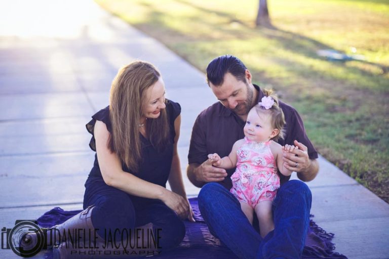 one year old birthday girl sitting on father's lap as mommy talks to her during golden hour at park on sidewalk captured by memphis photographer danielle jacqueline