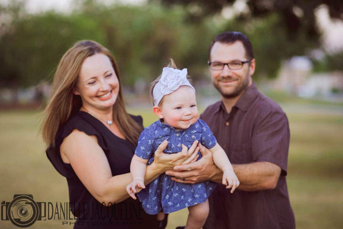 young parents at park during spring with 1 year old little girl in both of their arms extended towards camera with big smile for celebration portraits with danielle jacqueline photography servicing memphis tennessee