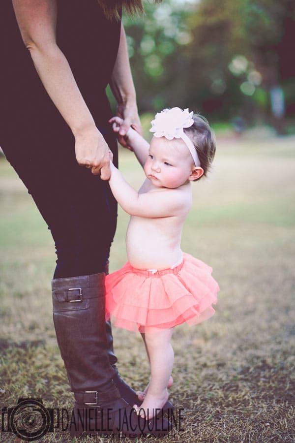 infant one year old girl in pink tutu with white bow standing on mother's feet eye level with child looking off to the right at park for creative session for memphis tn parents with danielle jacqueline photography