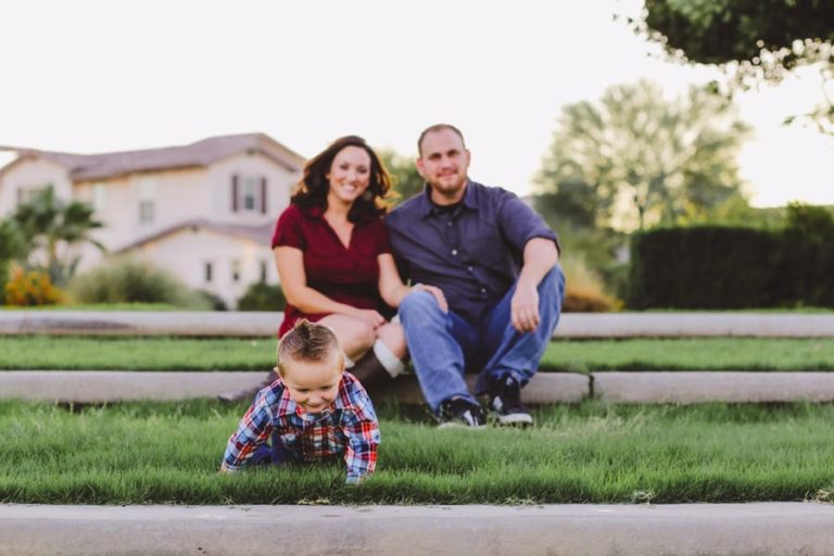 little toddler boy in plaid red white and blue shirt with hair spiked in grass while mom and dad sit on concrete steps onlooking cuddled up for fall family pictures with danielle jacqueline out of memphis tn