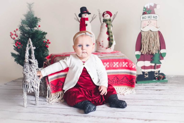 baby girl in red silk dress white wool sweater next to wicket reindeer faux pine tree and other christmas props during studio portrait session with danielle jacqueline photography in desoto county mississippi snowmen wood floor white edit