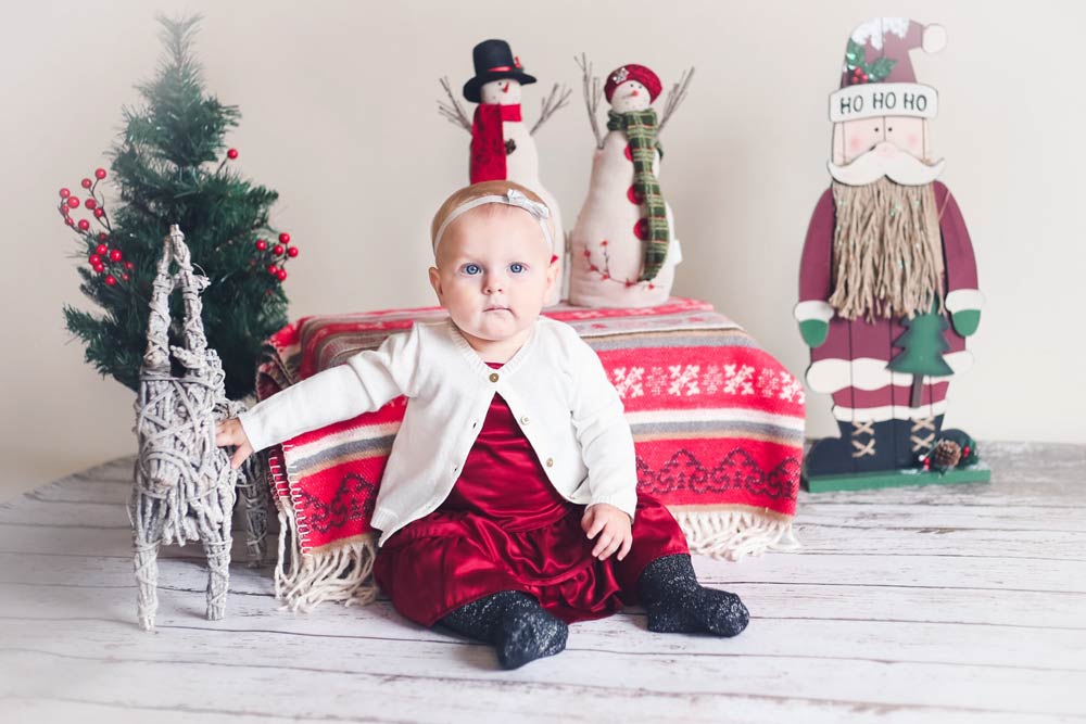 baby girl in red silk dress white wool sweater next to wicket reindeer faux pine tree and other christmas props during studio portrait session with danielle jacqueline photography in desoto county mississippi snowmen wood floor white edit