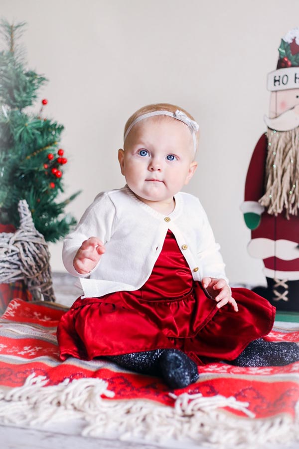 close up photograph of infant baby dressed in christmas colored outfit for family holiday pictures at studio with santa and evergreen tree props on woven blanket arm raised half smile soft glow memphis tn