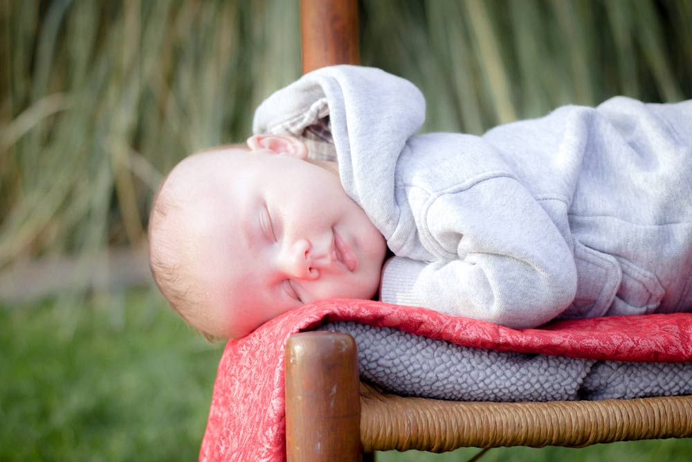 little boy in grey sweat suit asleep on brown wooden rocking chair with christmas colored blanket and green grass and vegetation during outdoor photo session with danielle jacqueline in olive branch ms