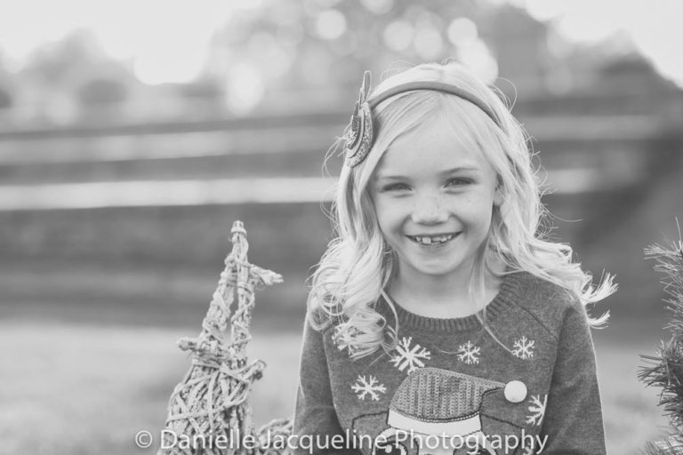 pre teen daughter of proud parents in desoto county ms sitting next to christmas props in front of camera smiling during outdoor photoshoot by danielle jacqueline sweater and bow in hair wicker reindeer black and white holiday portraits