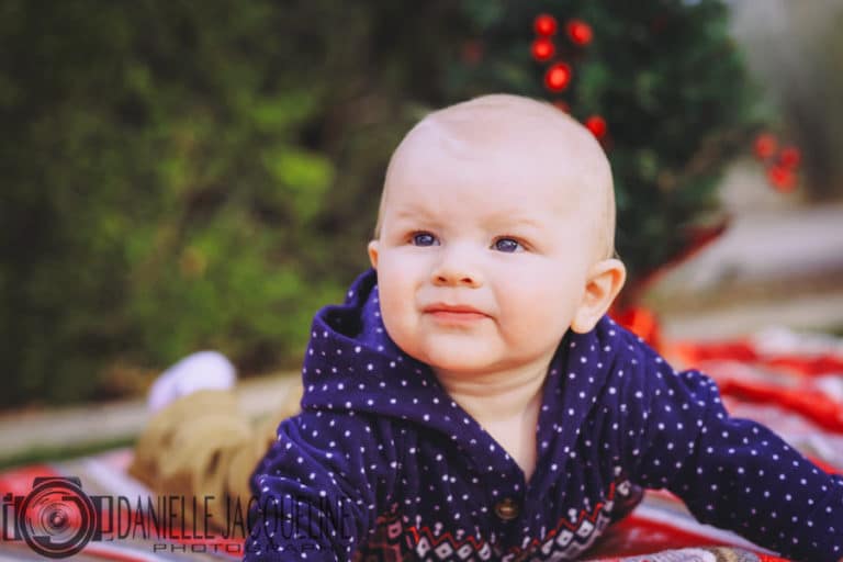 young laddy looking to his right while laying on chirstmas colored blanket in front of a fur pine tree in holiday sweater for family pictures with danielle jacqueline photography in olive branch ms