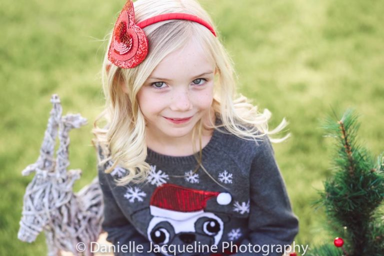 adorable children's photography by danielle jacqueline in desoto county looking down on blonde 9 year old daughter with red headband and how wearing grey christmas sweater with art in the middle of park for winter holiday cards