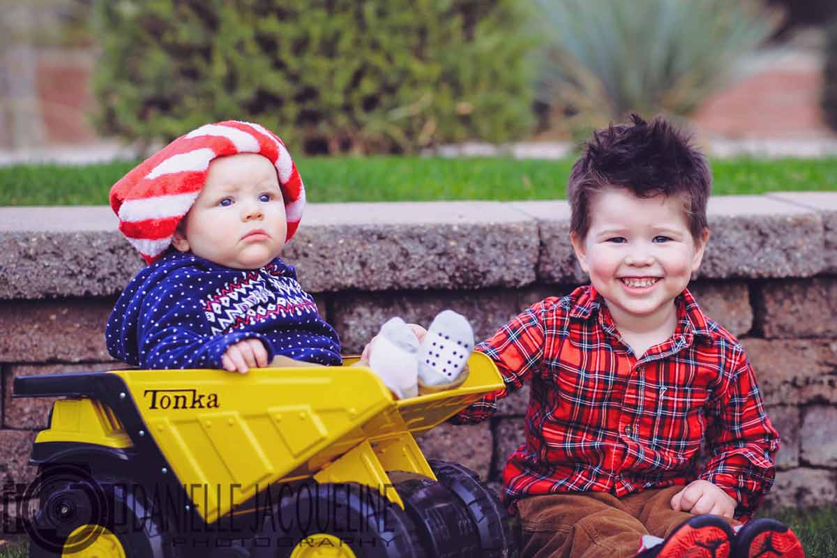 big brother with his new little sibling seated inside a big yellow tonka truck in front of stone slates with grass plateau outdoors for junior photo session with danielle jacqueline out of olive branch mississippi