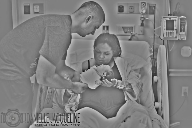 father handing off newly born baby girl to mother sitting in hospital recover bed propped up with pillows plugged into monitors during fresh 48 session with danielle jacqueline photography near memphis