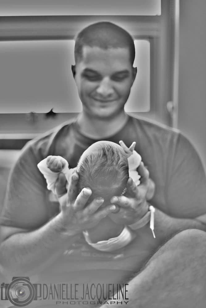 first time father smiling and looking down at his little infant daughter in his hands while sitting in hospital chair in recovery room with wife during fresh 48 shoot with danielle jacqueline photography in olive branch mississippi