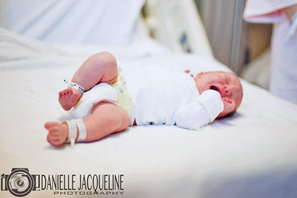 little baby at hospital after being born healthy to parents laying on linen sheets on bed crying with finger in mouth beanie on head fresh 48 session near memphis tn by danielle jacqueline photography