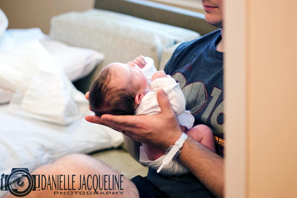 photograph of father holding newly born infant girl peeking around the corner capturing part of wall authentic intimate family moment for parents near memphis by photographer danielle jacqueline