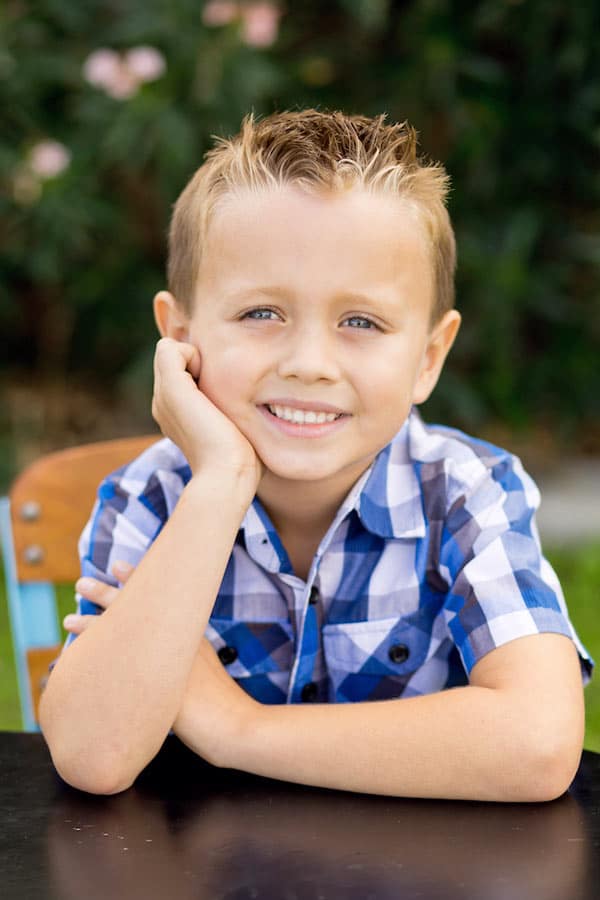 young boys palm to chin eyes straight ahead at lens centered on desk wearing blue and white checkered shirt in front of bushes in memphis tennessee by danielle jacqueline photography