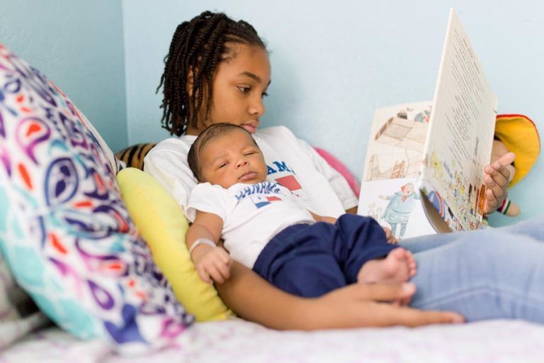 baby boy looking directly at camera lounging back on teal pillows while big sister elementary school is reading him a paperback story book before bedtime sibling photography by danielle jacqueline in olive branch ms south of memphis tn