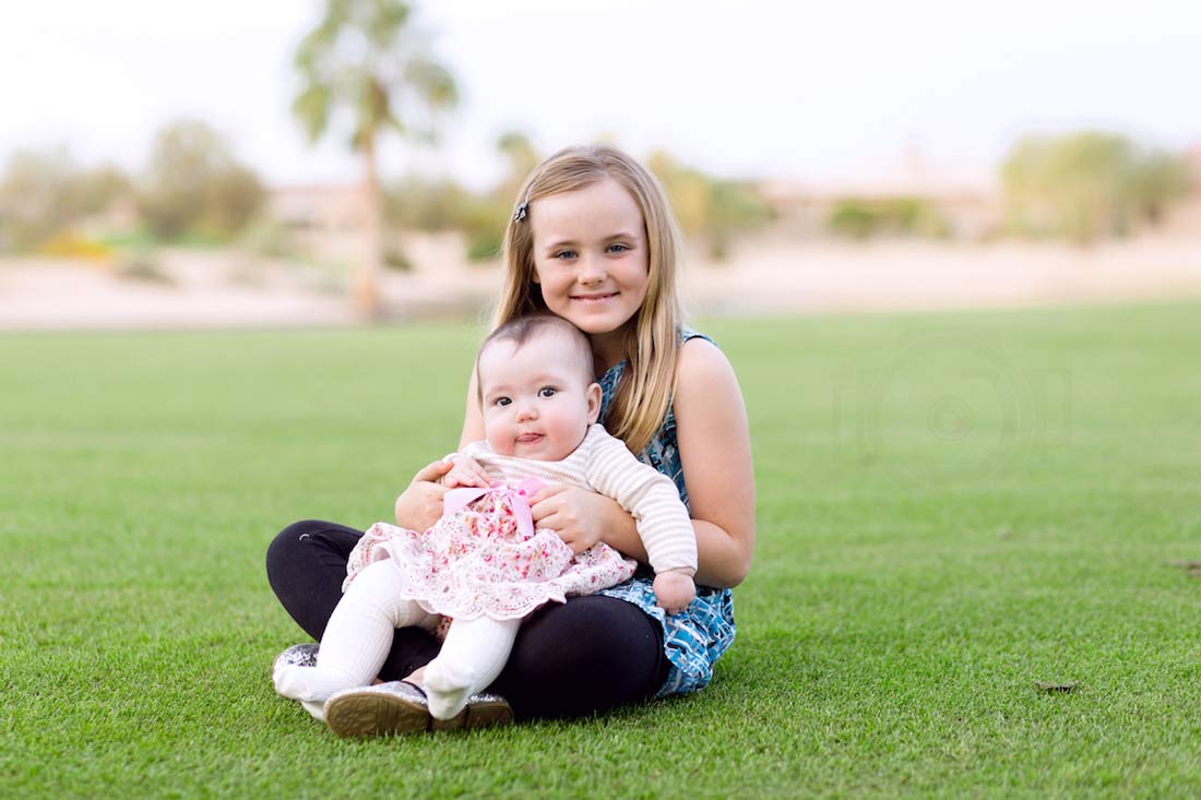 seated in grass in open field trees in back older sibling with baby sister on lap for family portrait session with danielle jacqueline photography just south of memphis tennessee