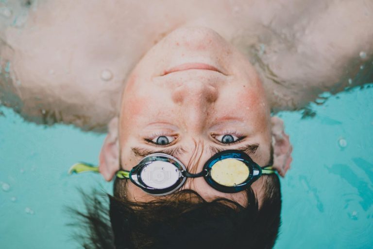 upside down portrait of high school senior boy in swimming pool with googles on forehead floating on back towards edge of deck where danielle jacqueline photography camera is near memphis tennessee